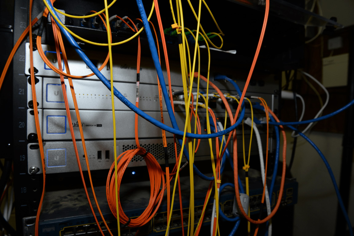 Dense bundle of orange, blue, and yellow network cables connecting switches in a server rack, representing the fiber and networking layer AI companies are racing to own