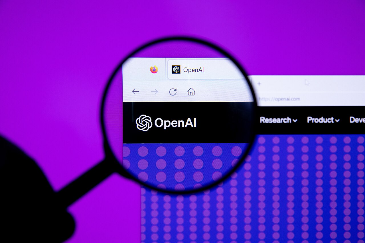 OpenAI website viewed through a magnifying glass, representing the GPT-5.6 government safety review