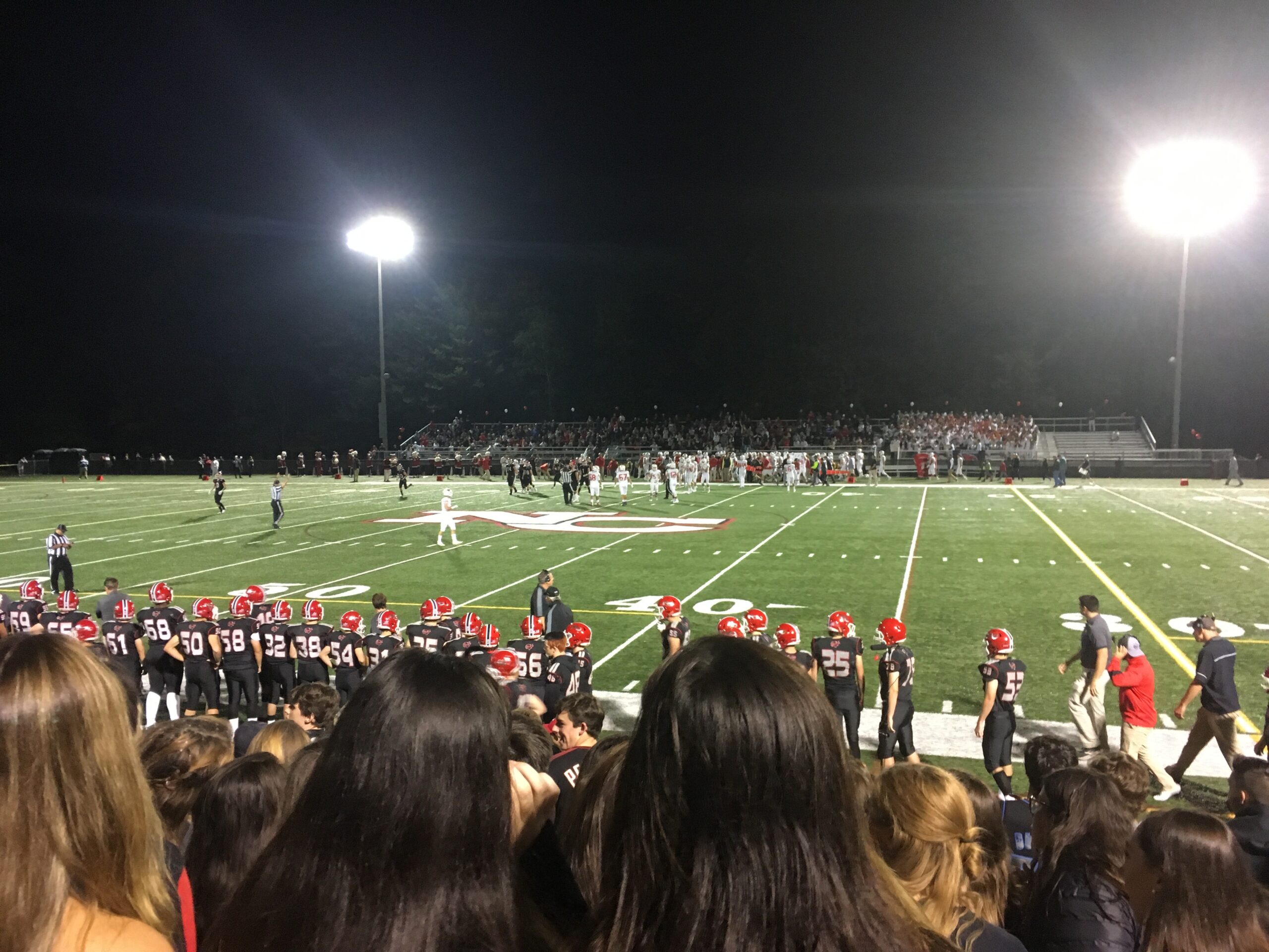 American football game under stadium lights showing players in red helmets and black uniforms lining up on the field with fans in the stands