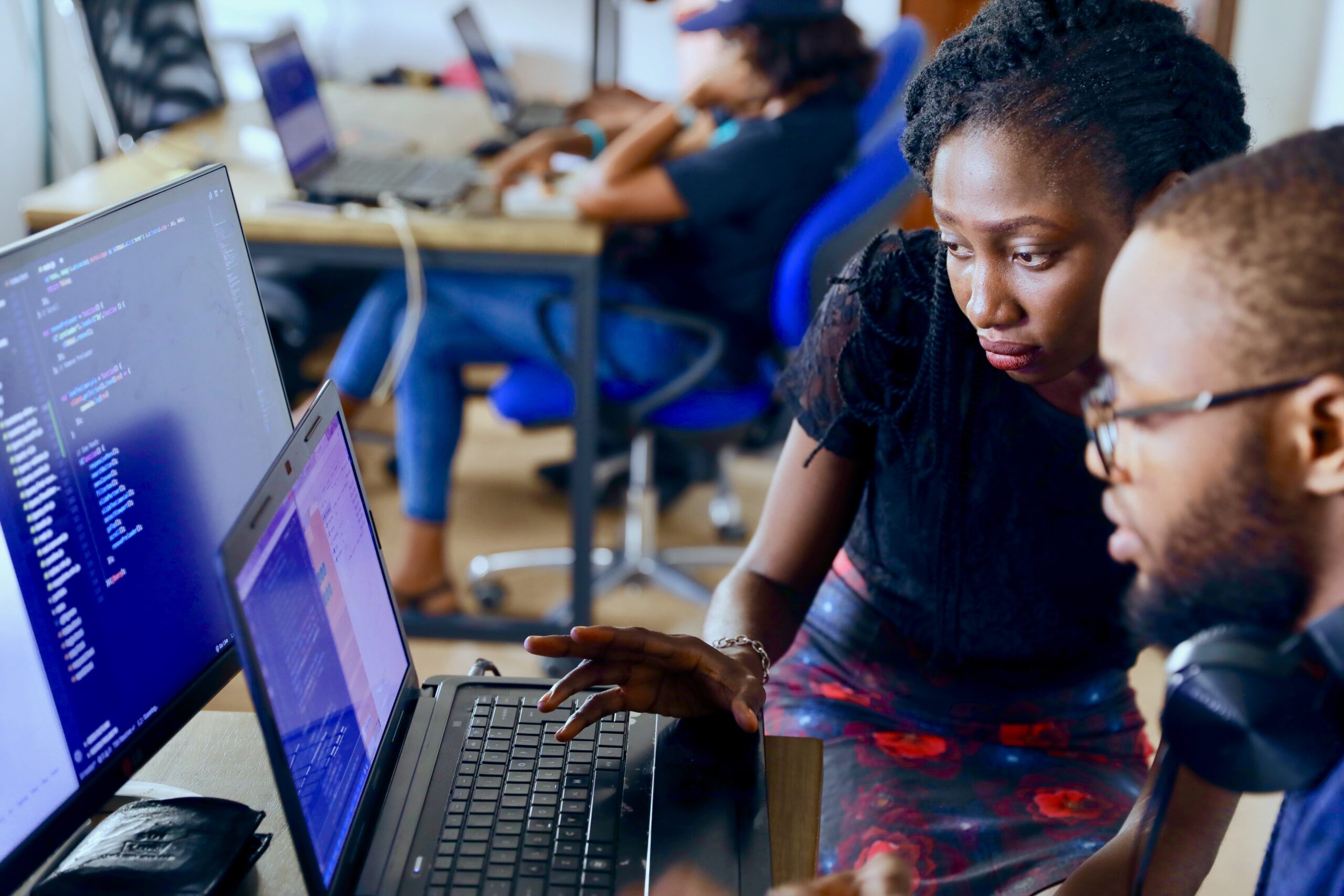 Two software developers collaborating at a laptop with colorful code visible on screen in a modern office environment with other programmers in the background
