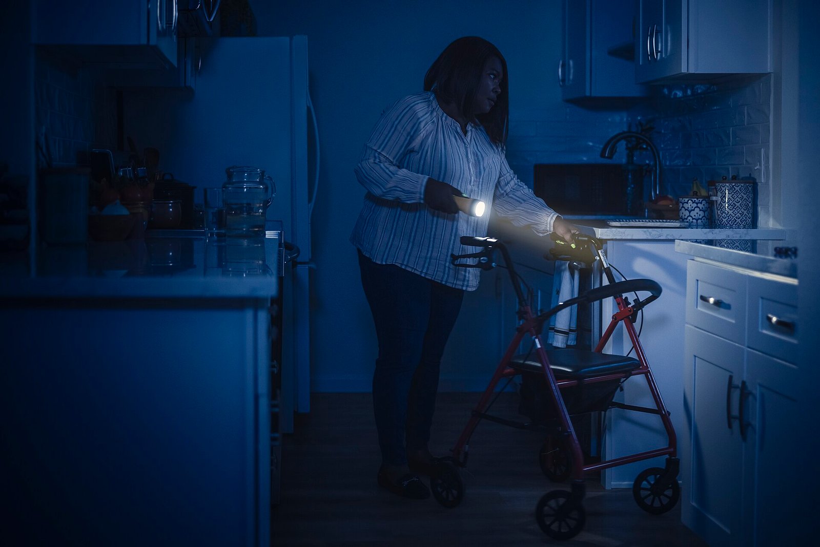 Person using a walker navigates a dark home kitchen with only a handheld flashlight during a power outage, illustrating why backup power matters during a blackout