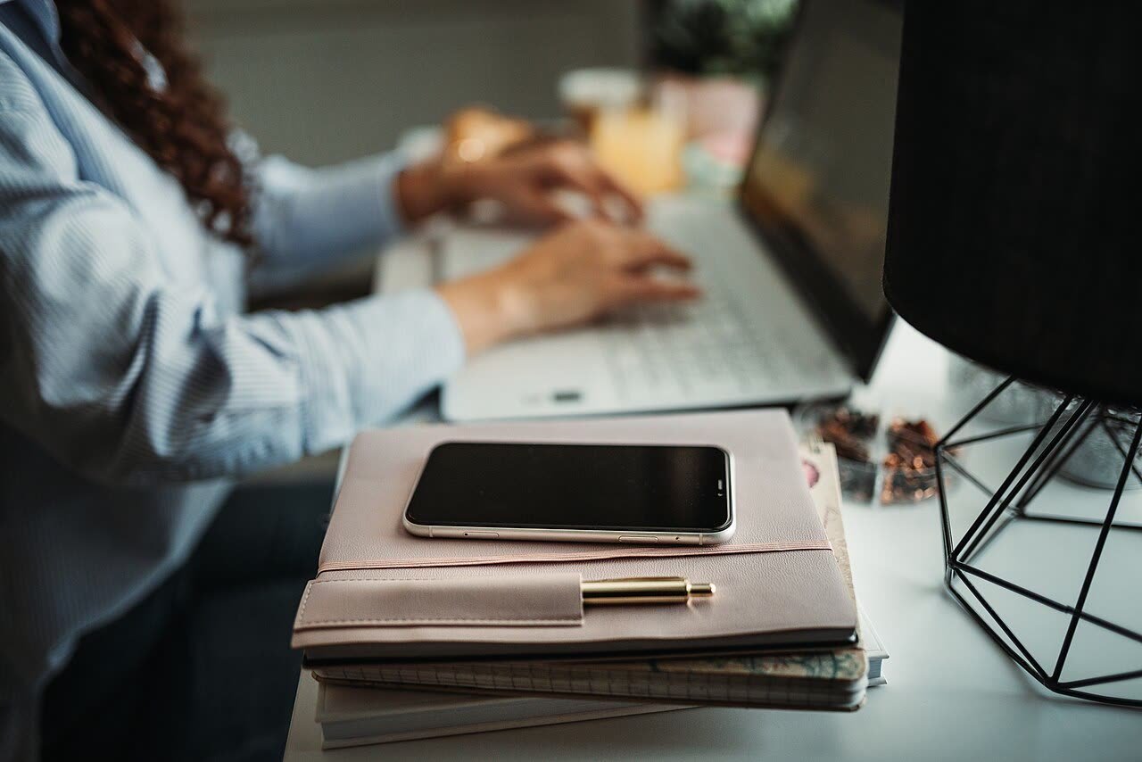 Person typing on a laptop with a paper planner and smartphone stacked nearby, symbolizing notes, tasks, and phone apps converging